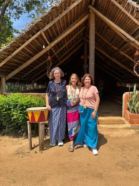 Three travelers wearing sarongs and wooden beads posing beside a colorful drum at a village hut
