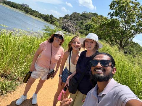 Group selfie of four smiling travelers on a lakeside dirt path with tall grass, water and hills in the background