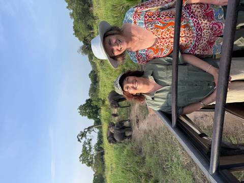 Two women in a safari jeep smile with a herd of wild elephants grazing in tall grass behind them