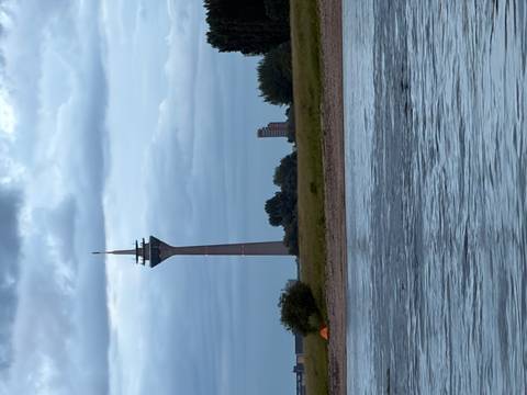 Tall telecommunications tower stands on a grassy riverbank under a moody cloudy sky, reflected in the Rhine