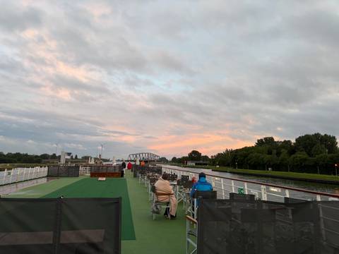 Passengers sit on deck chairs of a river cruise ship under a pastel evening sky with a distant bridge