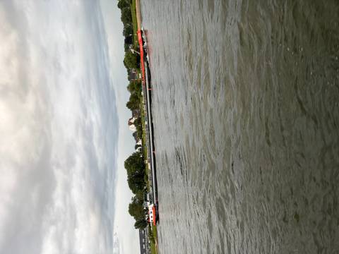 Long cargo barge travels along a wide river under a cloudy sky with shoreline houses behind