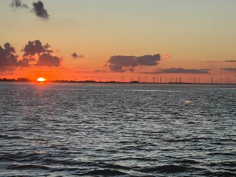 Vibrant orange sunset over open water with silhouettes of distant wind turbines and low clouds