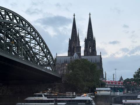 View of Cologne Cathedral’s twin spires beside the steel arches of Hohenzollern Bridge under a grey evening sky.