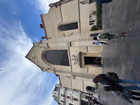 Stone church with a large arched window and clock above the entrance as visitors file through the doorway on a bright day.