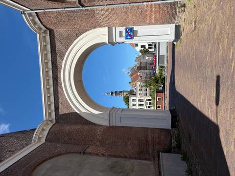 White stone archway framing a distant Dutch clock tower and traditional brick houses under clear blue sky.