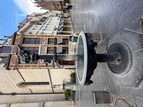Small ornate fountain topped by a bronze statue in a stone-paved city square.