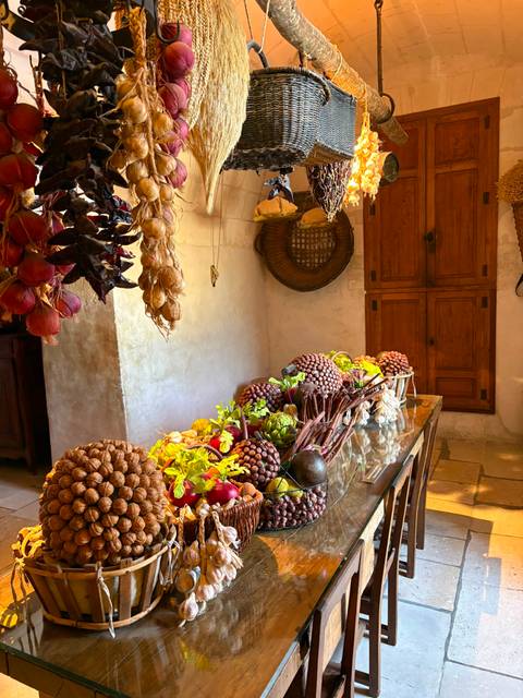 Rustic chateau kitchen with hanging onions and peppers, and a wooden table piled with colourful fruits, nuts and vegetables.