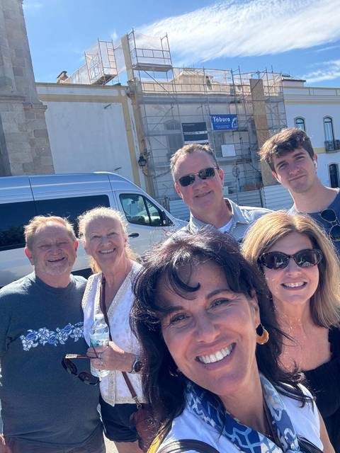 Cheerful selfie of six travellers smiling in bright sun with a tour van behind them