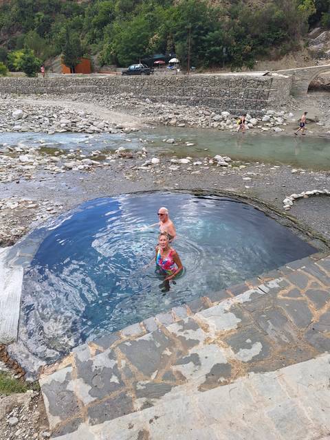 Two seniors enjoy a soak in a natural circular hot-spring pool beside a rocky river.