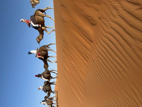 Line of riders on camels crest a golden dune against a deep blue desert sky.