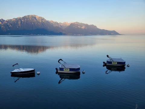 Three small motorboats floating on a calm lake reflecting distant alpine peaks at dawn.
