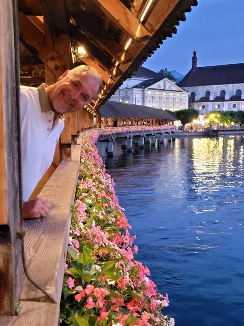 Man leaning from flower-lined wooden chapel bridge over the Reuss River at dusk.