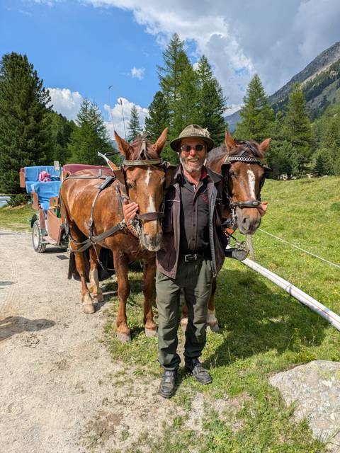 Coachman cheerfully standing between two harnessed carriage horses on a mountain trail.