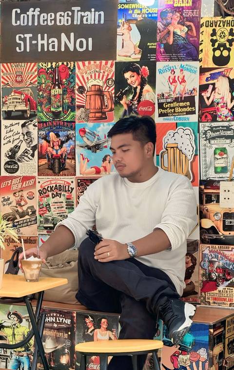 Young man in white sweater sitting at a cafe table in front of colorful retro posters.