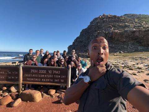 Excited tour group selfie at Cape of Good Hope sign with rocky coastline and blue ocean backdrop.