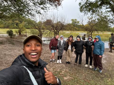 Rain-soaked travellers posing in ponchos beside their smiling guide amid a flooded woodland campsite.