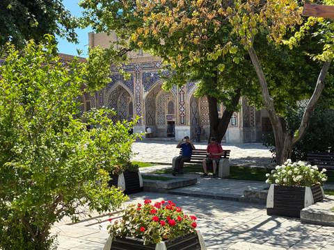 Leafy courtyard of a decorated madrasah with intricate blue tiling where two men rest on benches.