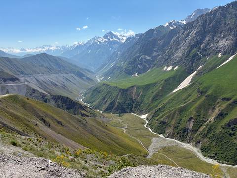 Sweeping alpine valley with a winding river, lush green slopes and distant snow-capped peaks under a bright blue sky.