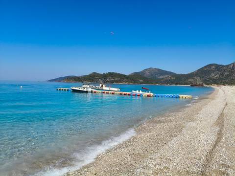 Crystal-clear turquoise sea laps a pebbly beach with moored speedboats and distant hills.