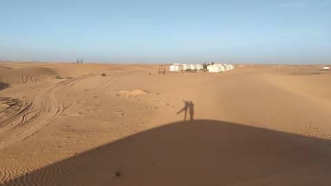 Remote desert camp of white tents in rolling dunes under a pale morning sky with photographer’s shadow.