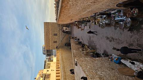 Market alley along fortified stone walls with stalls selling baskets and goods; locals and tourists strolling.