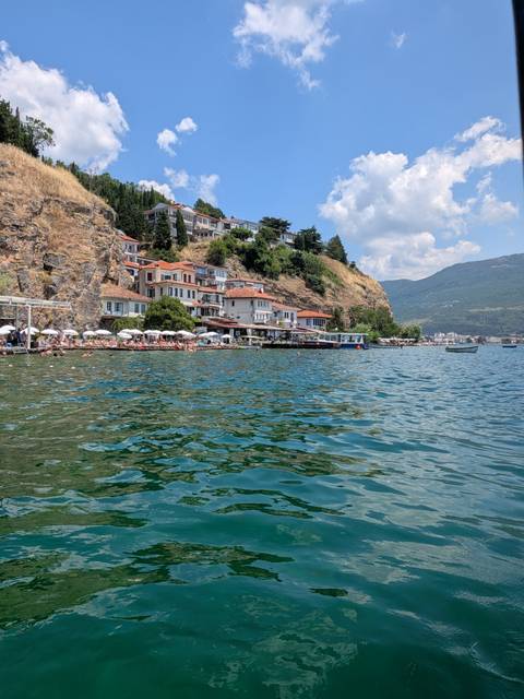 Waterside restaurants and sunbathers along Lake Ohrid with rocky hill and mountains beyond.