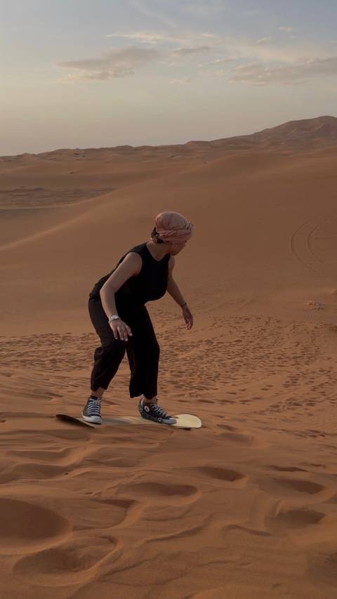 Woman in black outfit and headwrap balances on a steep orange sand dune.