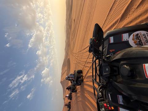 Several riders drive quad bikes across vast orange sand dunes under a blue sky dotted with thin clouds.