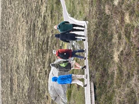 Hikers bundled in jackets walk along a wooden boardwalk across open heathland.
