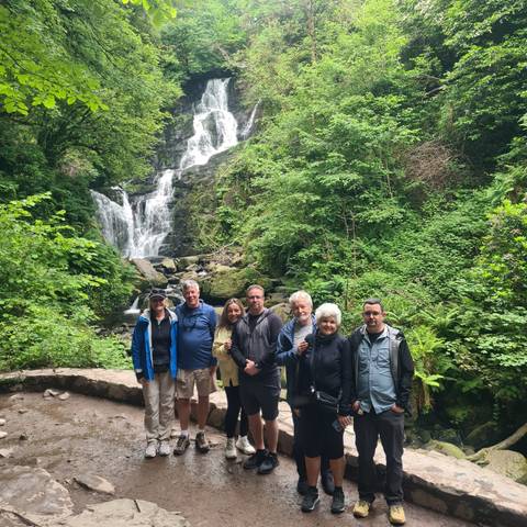 Smiling travellers stand before a lush forest waterfall surrounded by green foliage.