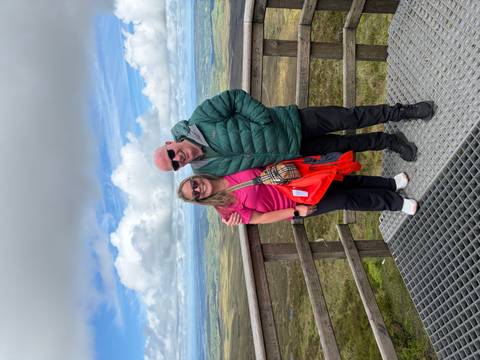 Smiling couple on a wooden viewing platform high above rolling Irish countryside under scattered clouds.