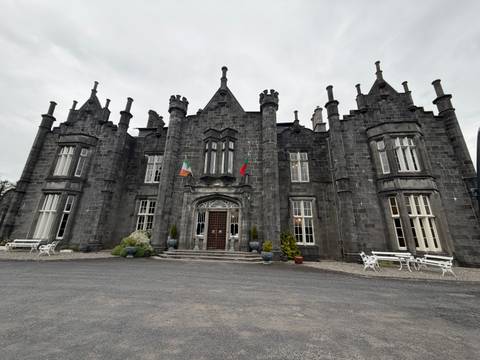 Grey stone Gothic-style manor with Irish and Portuguese flags, white benches and cloudy sky.