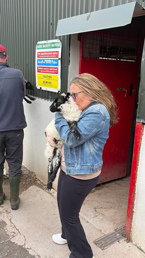 Woman in denim jacket cuddles a black-and-white lamb outside a farm building.