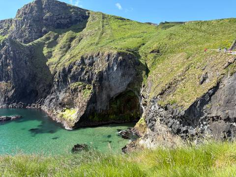 Crystal-clear turquoise cove and jagged sea cliffs near a rope bridge path with tiny hikers visible.