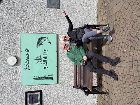 Cheerful couple reclines on a bench beneath a mint-green "Welcome to Bushmills" sign.