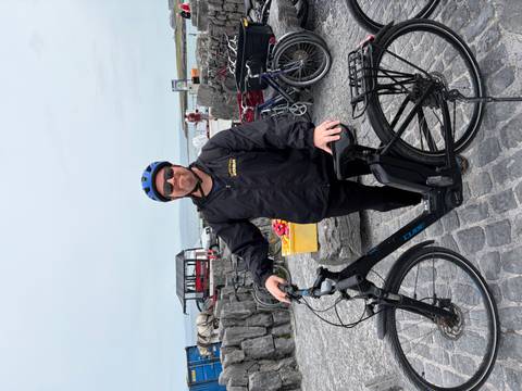 Cyclist wearing helmet poses with an e-bike near stone walls and horse carts by the sea.