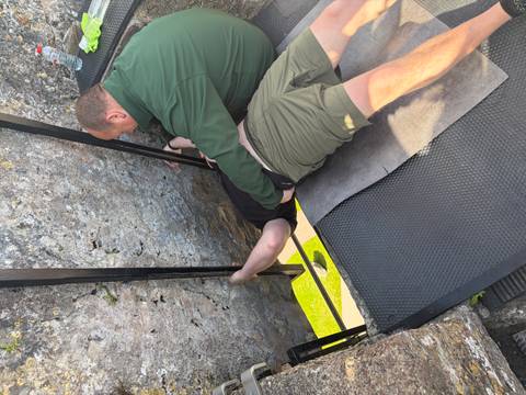 Visitor bends backward to kiss the Blarney Stone while a helper supports him on the castle parapet.
