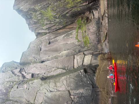 Red kayaks float on dark coastal waters beneath towering striated rock cliffs.