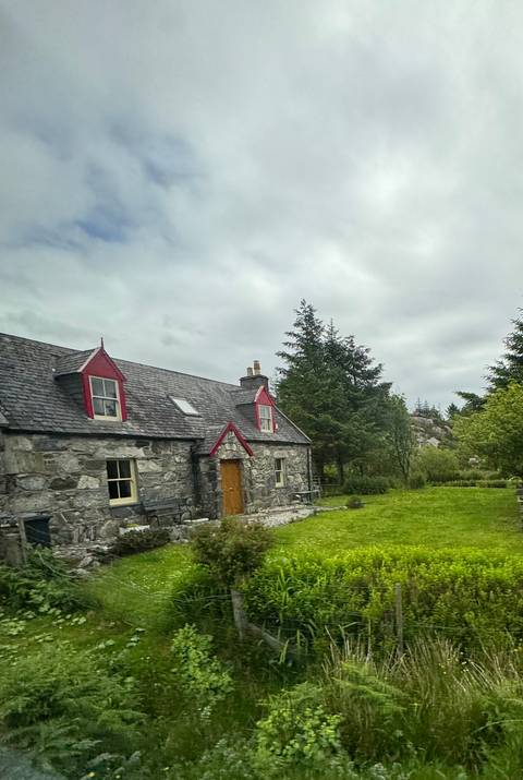A rustic stone cottage with red-trimmed dormers surrounded by evergreens under cloudy skies.