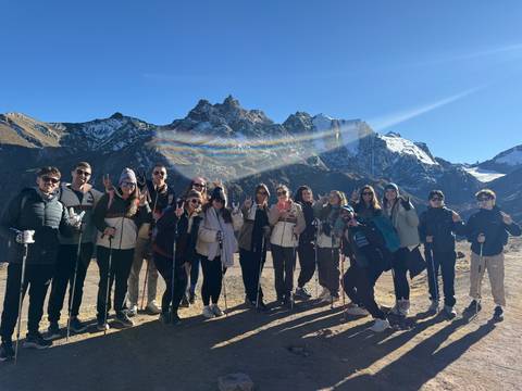 Large hiking group poses against snow-capped Andean peaks with a rainbow light flare arching overhead.
