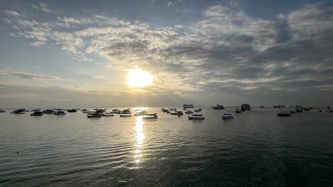Wide view of dozens of boats floating on tranquil sea water beneath a glowing low sun and scattered clouds.