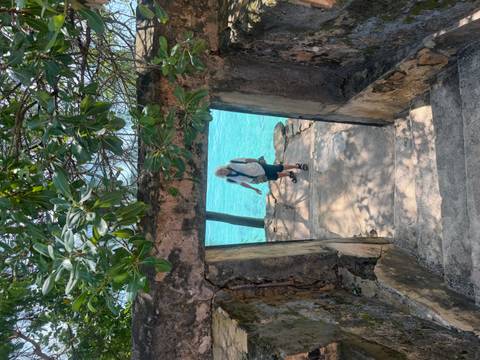 Traveler descends stone steps through a doorway toward bright turquoise sea framed by leafy branches.
