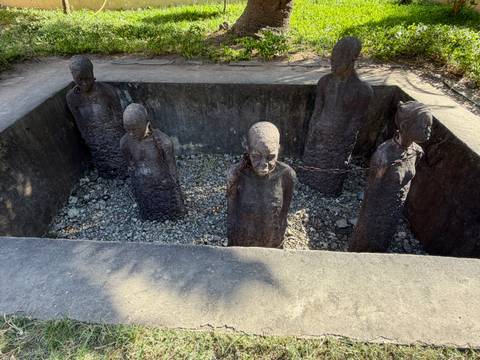 Stone statues of chained slaves stand in a sunken memorial pit, lit by late afternoon sun.