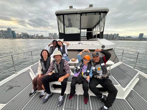 Happy group sitting on the bow of a yacht on a city harbour with modern skyline and grey skies.