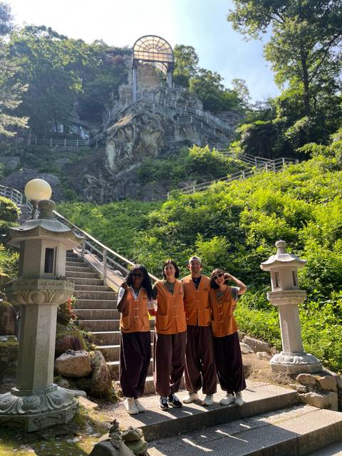 Four volunteers in orange vests stand at the base of a forested temple staircase on a sunny day.
