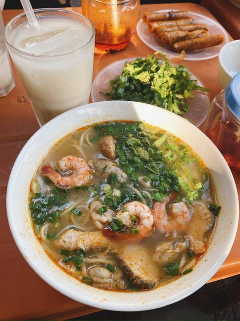 Bowl of aromatic shrimp noodle soup garnished with fresh herbs alongside iced drink on a plastic table.