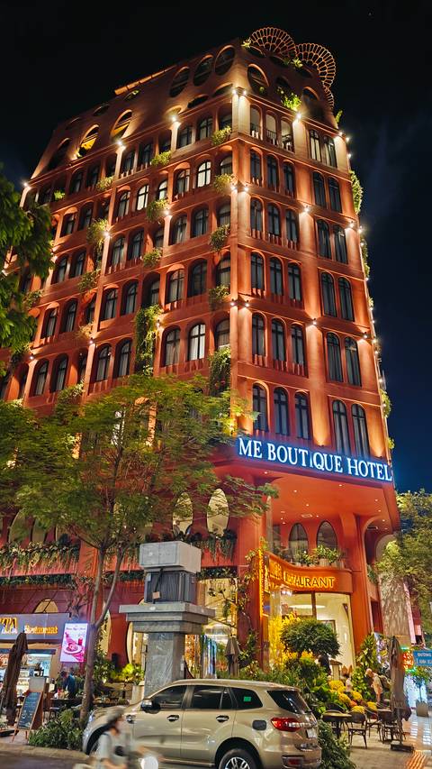 Night view of the illuminated red-orange façade of ME Boutique Hotel framed by leafy trees.