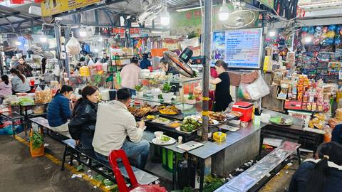 Bustling indoor food market with diners seated at narrow stalls surrounded by colorful packaged goods and menus.