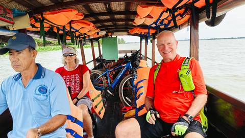 Cyclists and boat crew sitting in a wooden long-tail boat with life jackets and bikes onboard crossing a wide river.